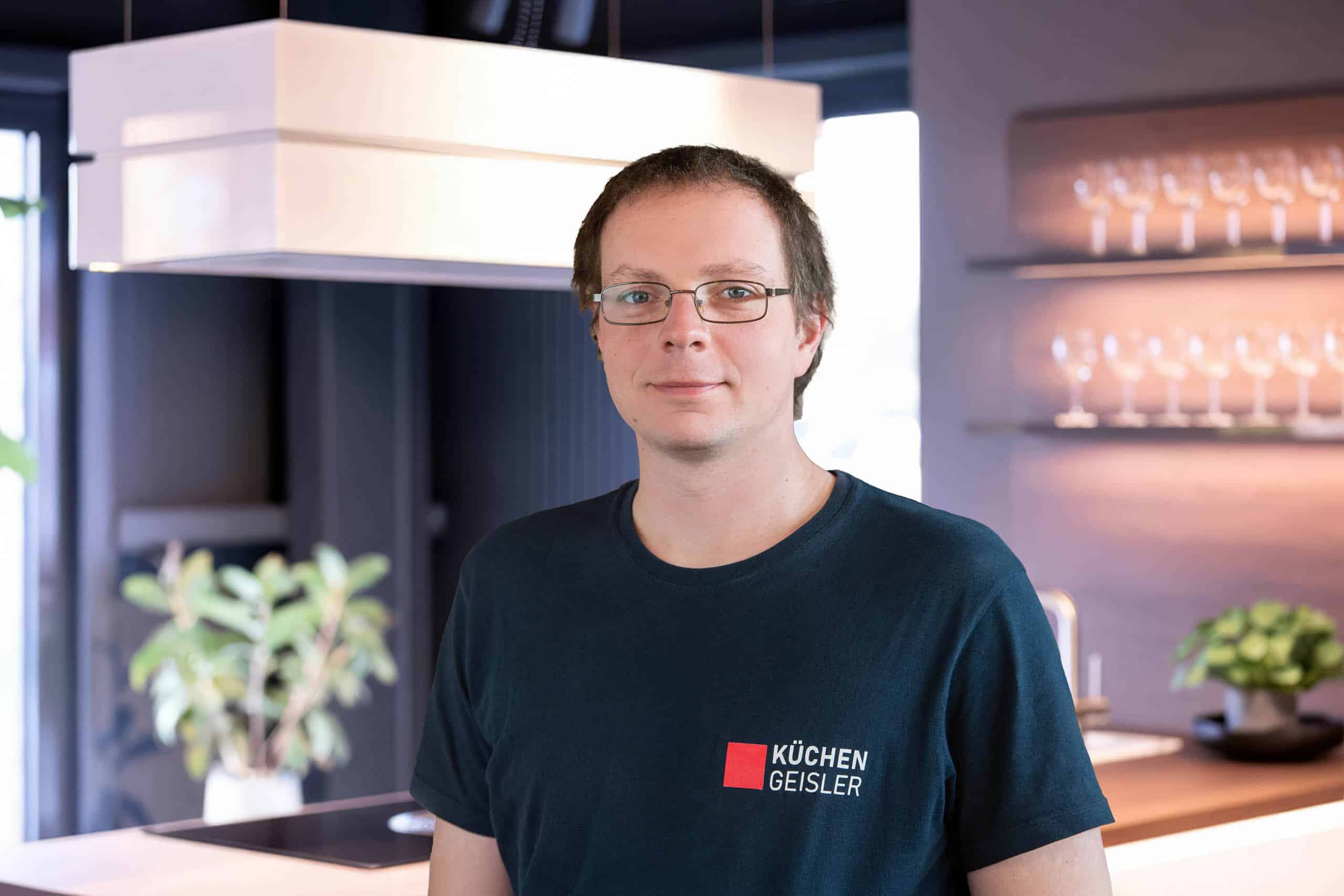 Ein Mann mit Brille und kurzen braunen Haaren steht in einer modernen Küche und trägt ein dunkles Hemd mit einem Küchen Geisler-Logo. Im Hintergrund sind Regale mit Glaswaren, Pflanzen und eine Küchenhaube zu sehen.