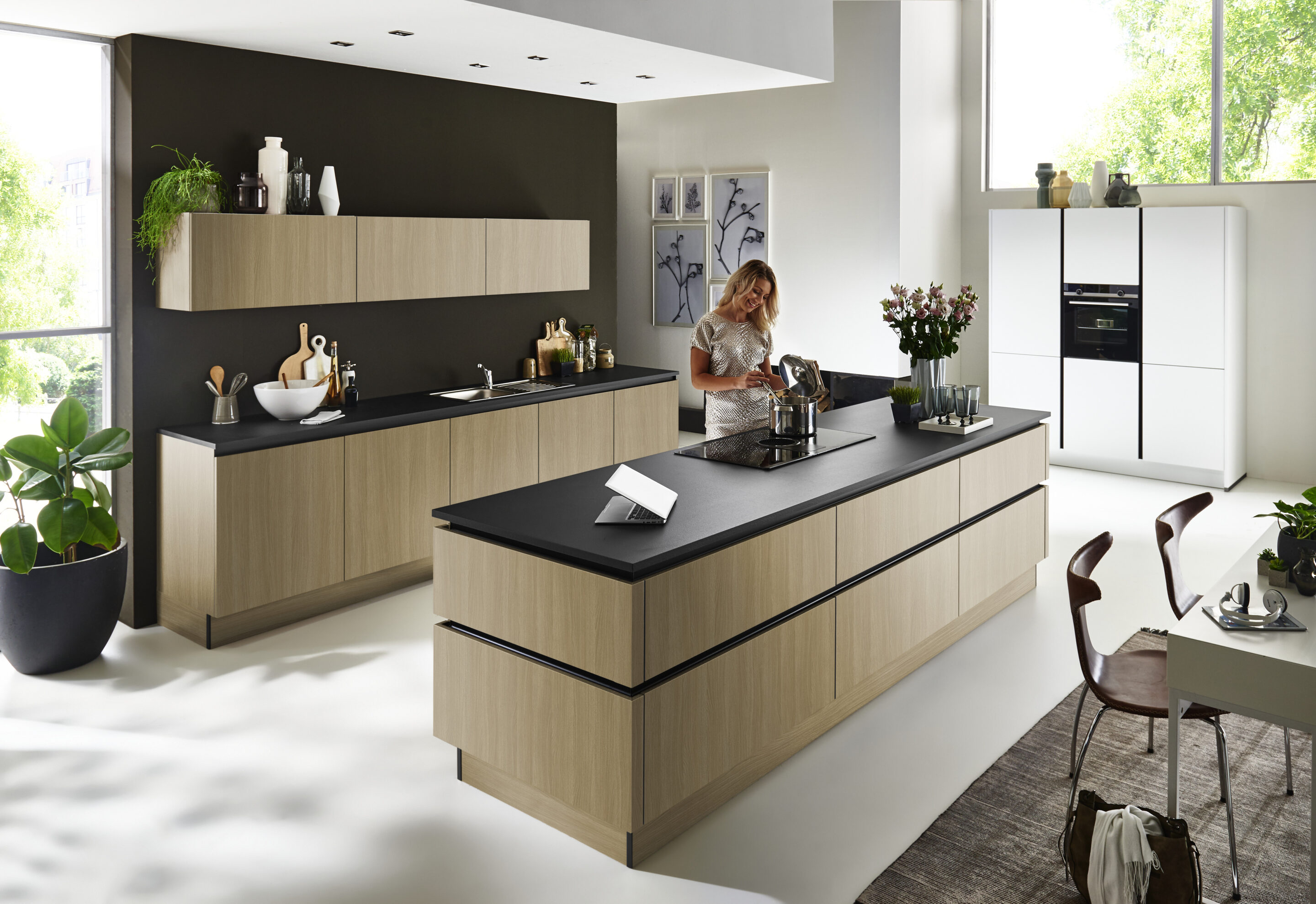 Eine Frau bereitet in einer modernen Küche mit hellen Holzschränken, schwarzen Arbeitsplatten, einer Kücheninsel mit cleveren Stauraumlösungen, Zierpflanzen und großen Fenstern, durch die Tageslicht hereinströmt, das Essen zu.